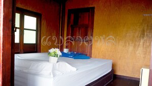 A neatly made bed with white linens, a small vase with flowers, and a blue cloth on a wooden platform in a room with wooden walls and a wooden door. - Saichol Floating Resort (Ban Ta Khun)