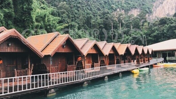 A row of wooden cabins with a white railing, connected by a bridge over a body of water, surrounded by dense greenery. - Saichol Floating Resort (Ban Ta Khun)