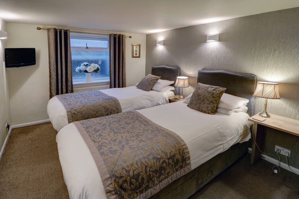 Una habitación de hotel con dos camas, televisión, ventana con cortinas y mesita de noche con lámpara.