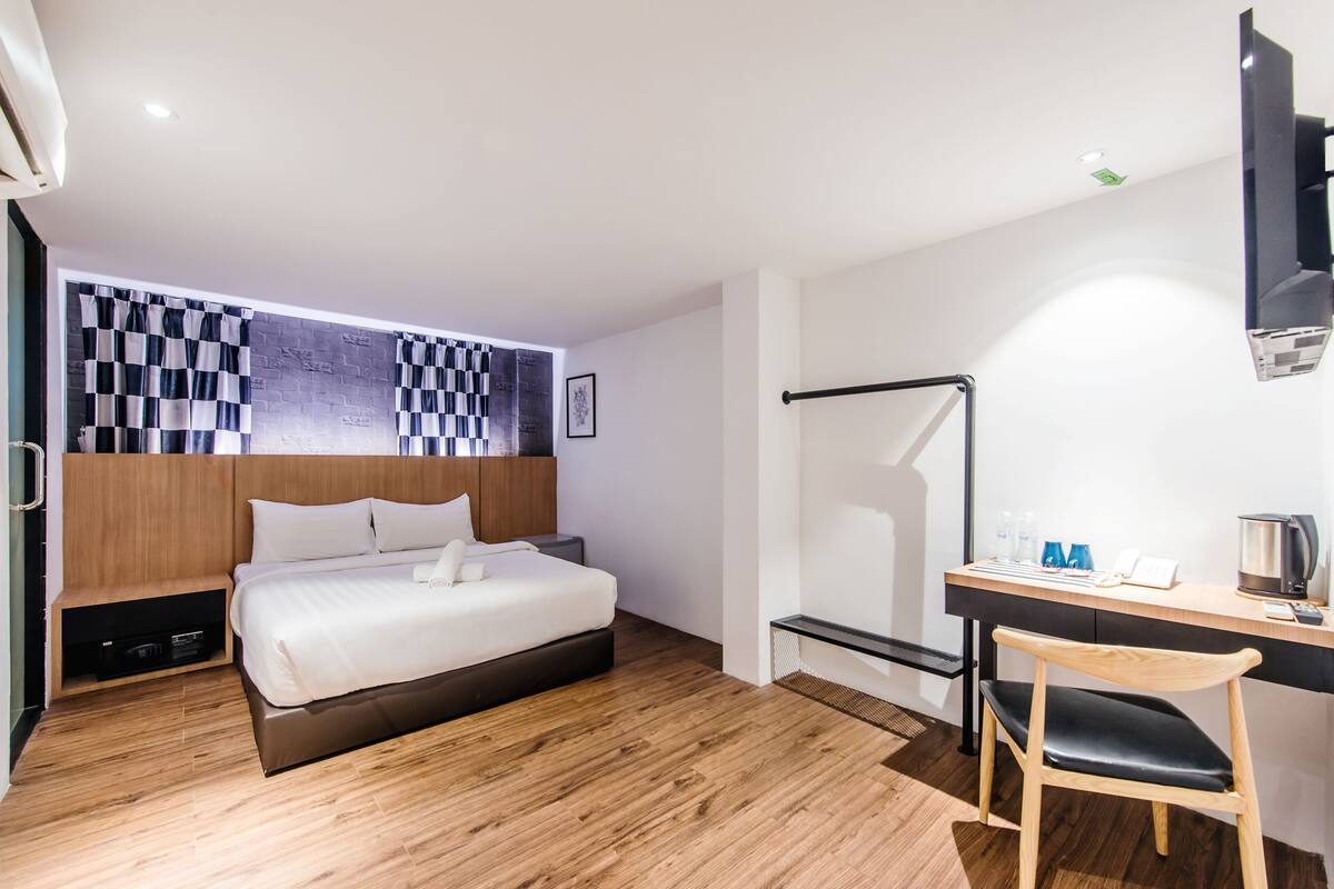 A modern hotel room with a wooden floor, a bed with white linens, a desk with a chair, a flat-screen TV, and a shower area.