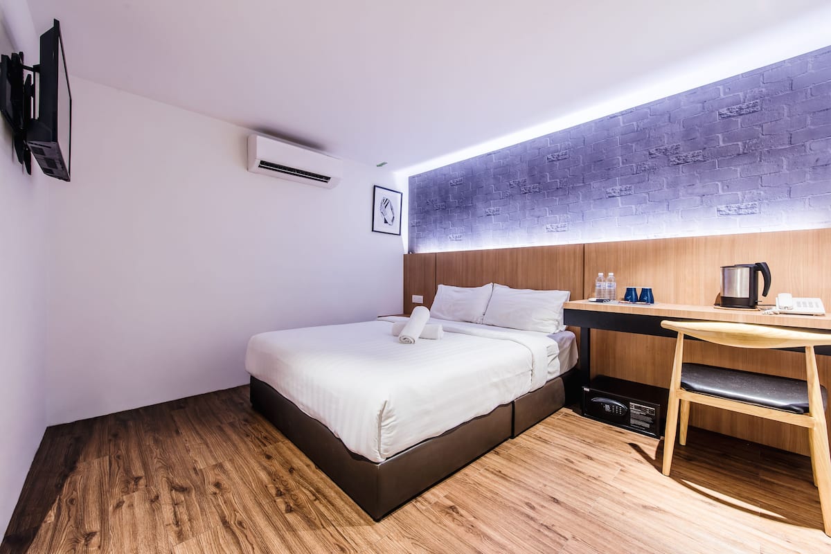 A modern hotel room with a wooden floor, a bed with white linens, a desk with a kettle, and a brick-patterned wall.