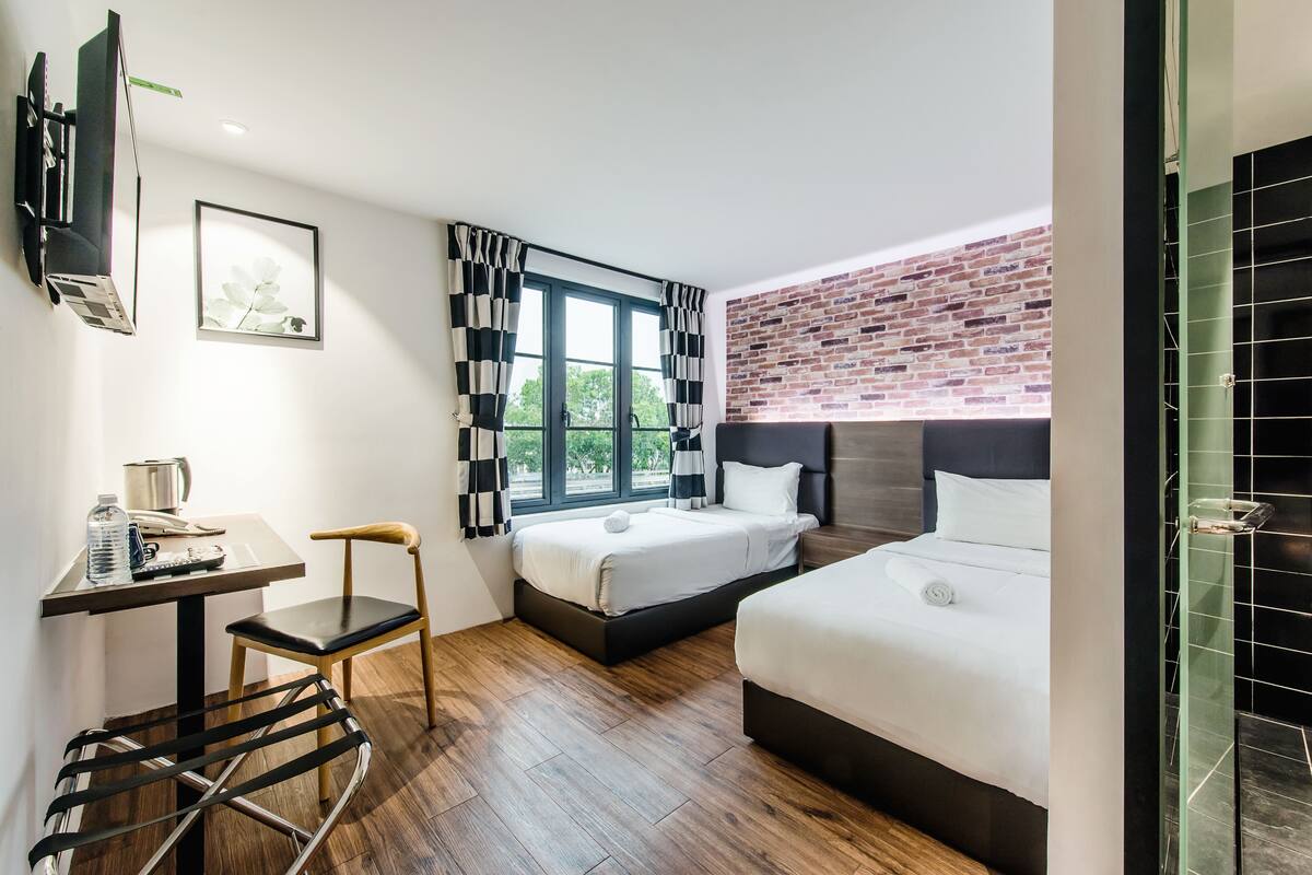 A hotel room with a desk, two beds, a chair, a window with curtains, a brick wall, and a shower area.