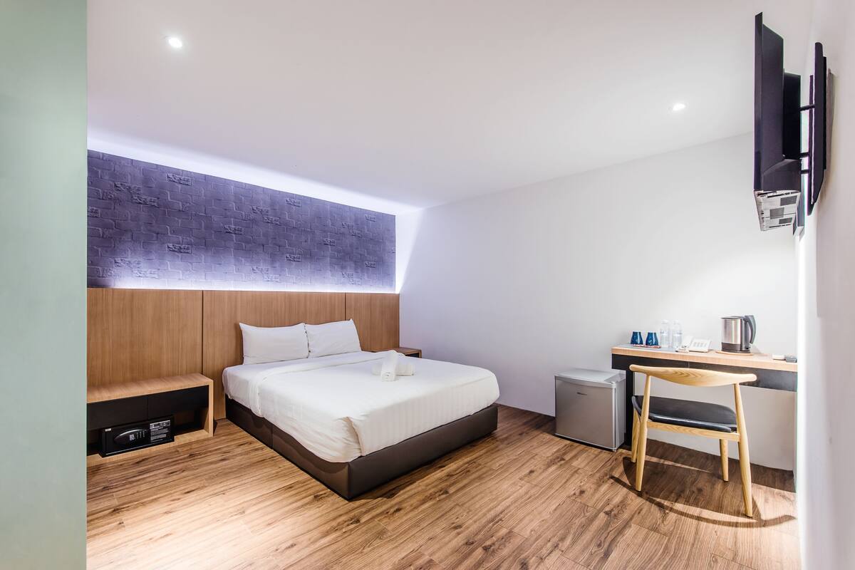 A modern hotel room with a wooden bed, a desk with a chair, a small refrigerator, and a flat-screen TV mounted on the wall.