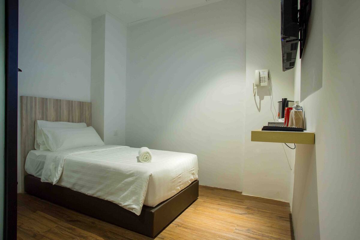 A neatly made bed with a white comforter and pillows, a wooden headboard, a bedside table with a lamp, and a mounted television.
