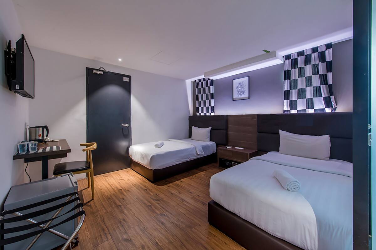 A hotel room with two beds, a desk, a chair, a small table with a kettle, and a checkered curtain.