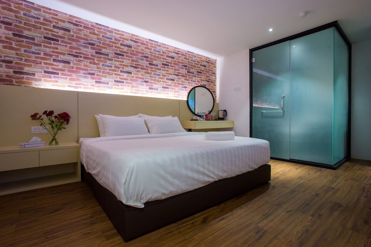 A modern hotel room with a brick-patterned wall, a large bed with white linens, a bedside table with a vase of flowers, a mirror, and a wardrobe with frosted glass doors.