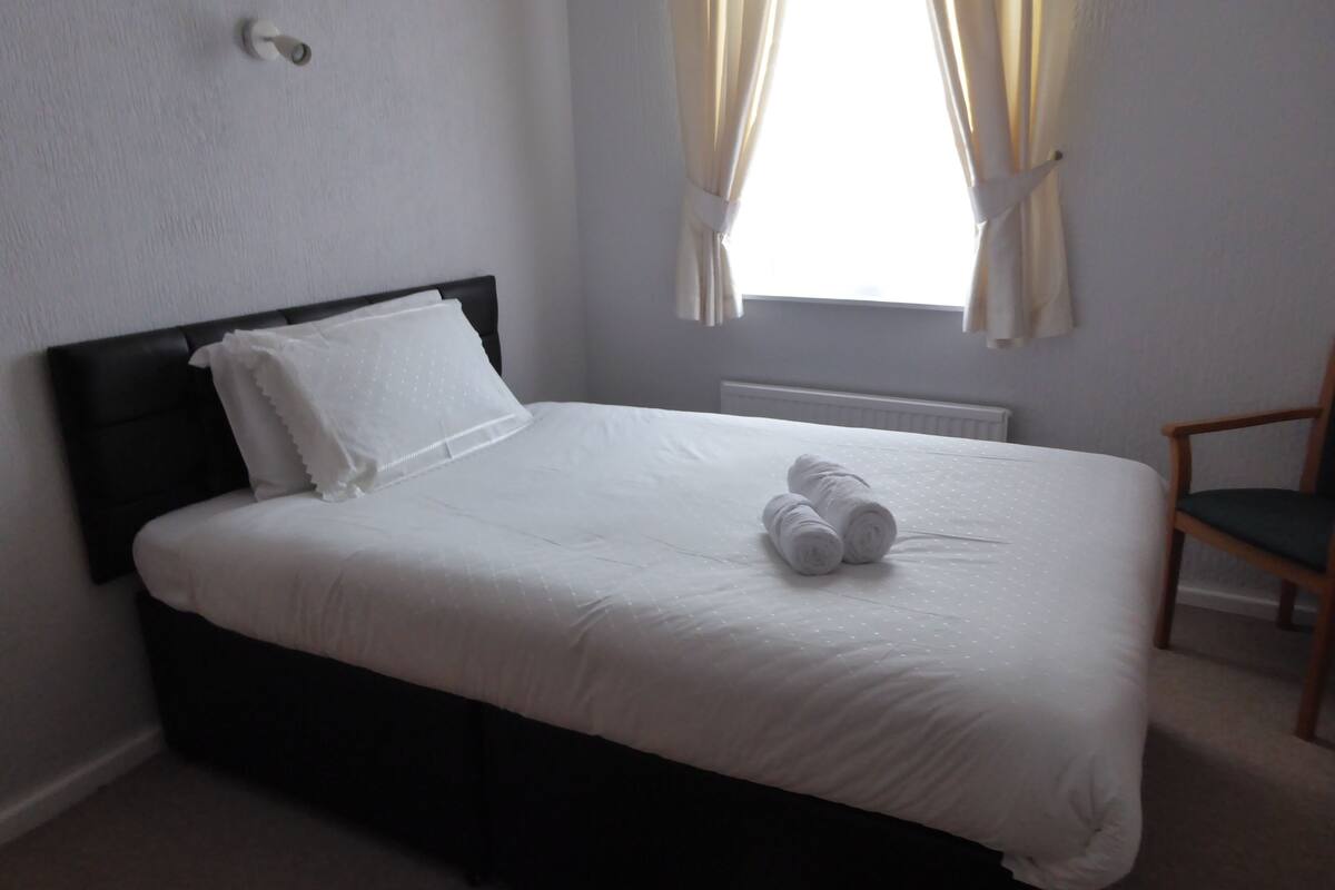 A neatly made bed with white linens and two rolled towels, a wooden headboard, a wooden chair, and a window with curtains.