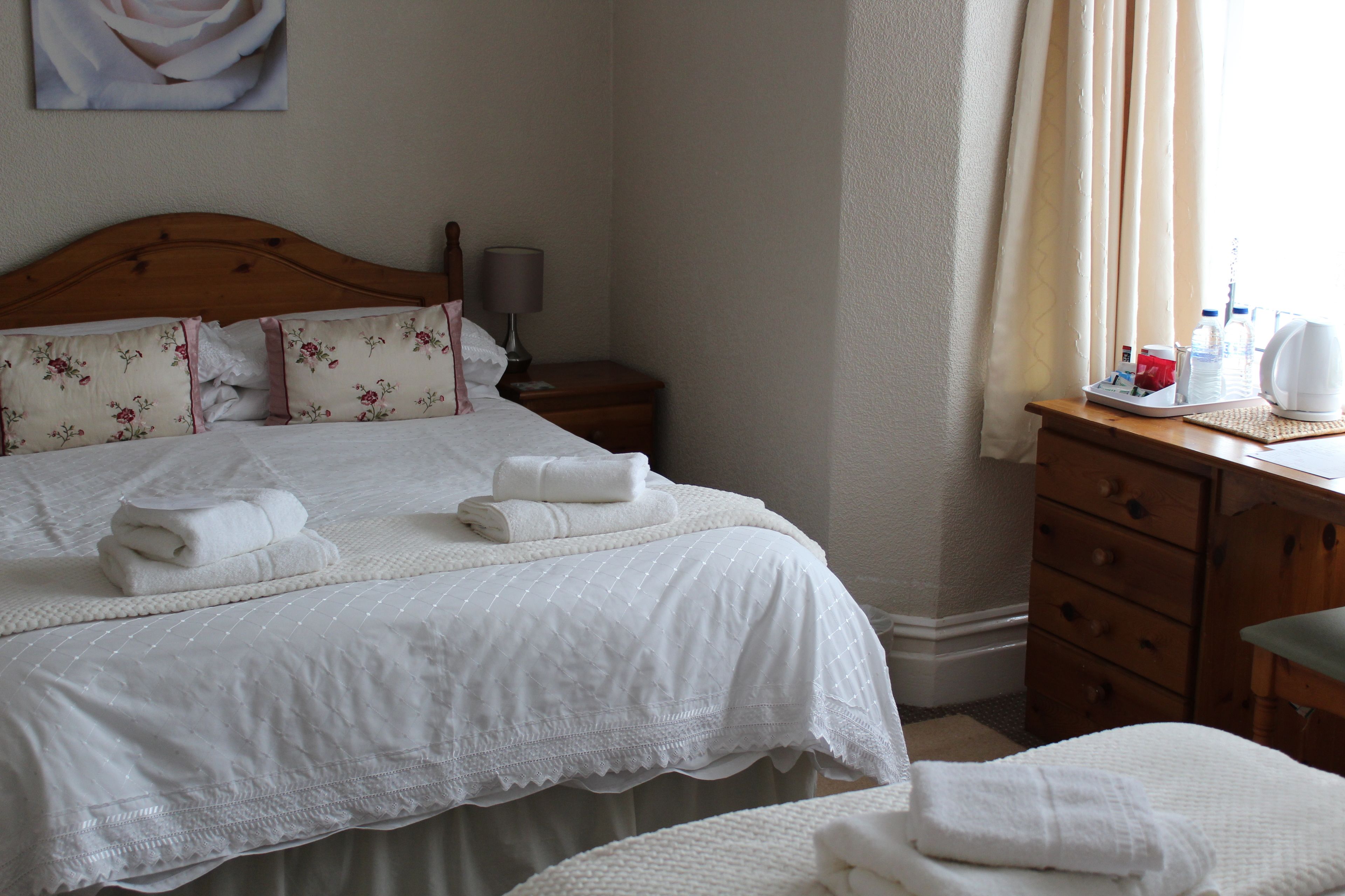 A neatly made bed with white linens and a floral pillow, a wooden nightstand with a lamp, and a wooden dresser with toiletries.