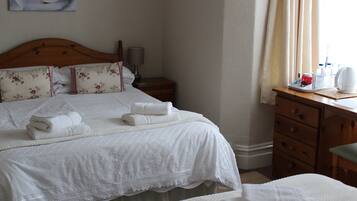 A neatly made bed with white linens and a floral pillow, a wooden nightstand with a lamp, and a wooden dresser with toiletries.