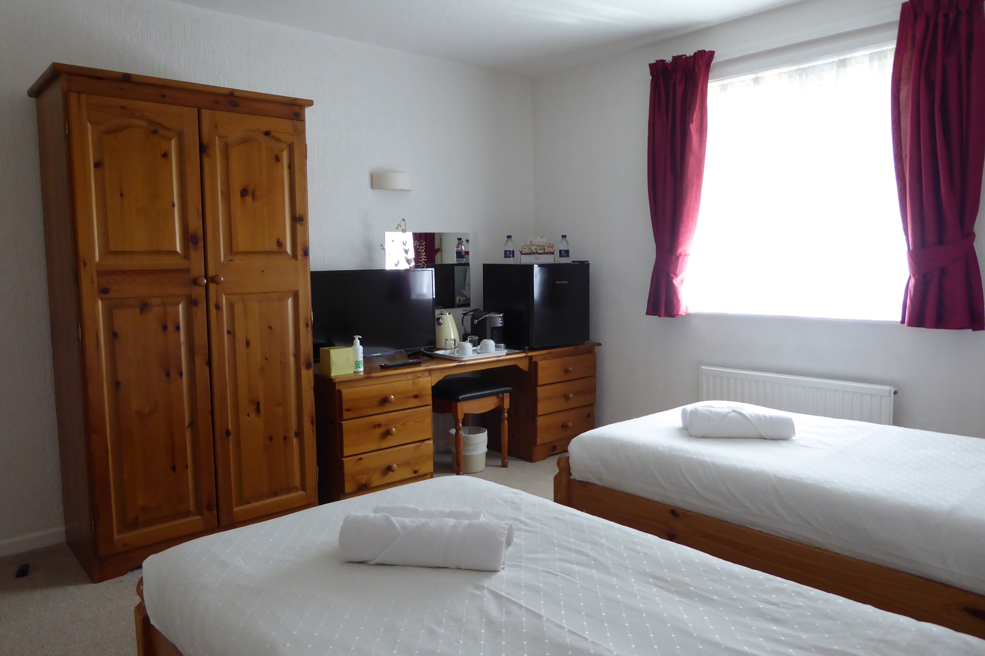 Quarto com duas camas, um guarda-roupa de madeira, uma mesa com computador e uma janela com cortinas.