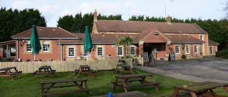 A brick building with a tiled roof, outdoor seating with picnic tables and umbrellas, and a paved entrance.