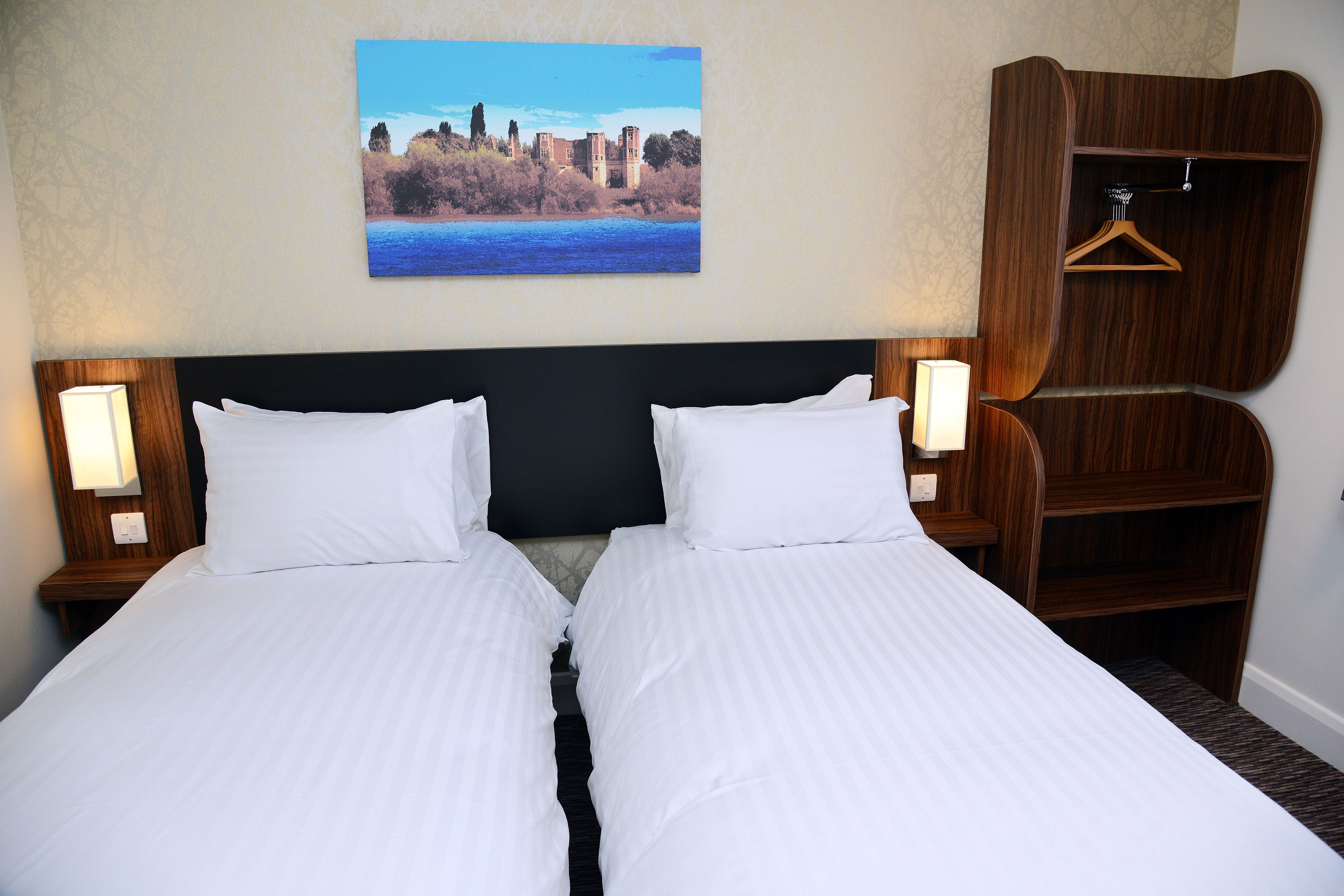 Quarto de hotel com duas camas, cabeceira de madeira, pintura de uma paisagem e guarda-roupa de madeira com cabides.