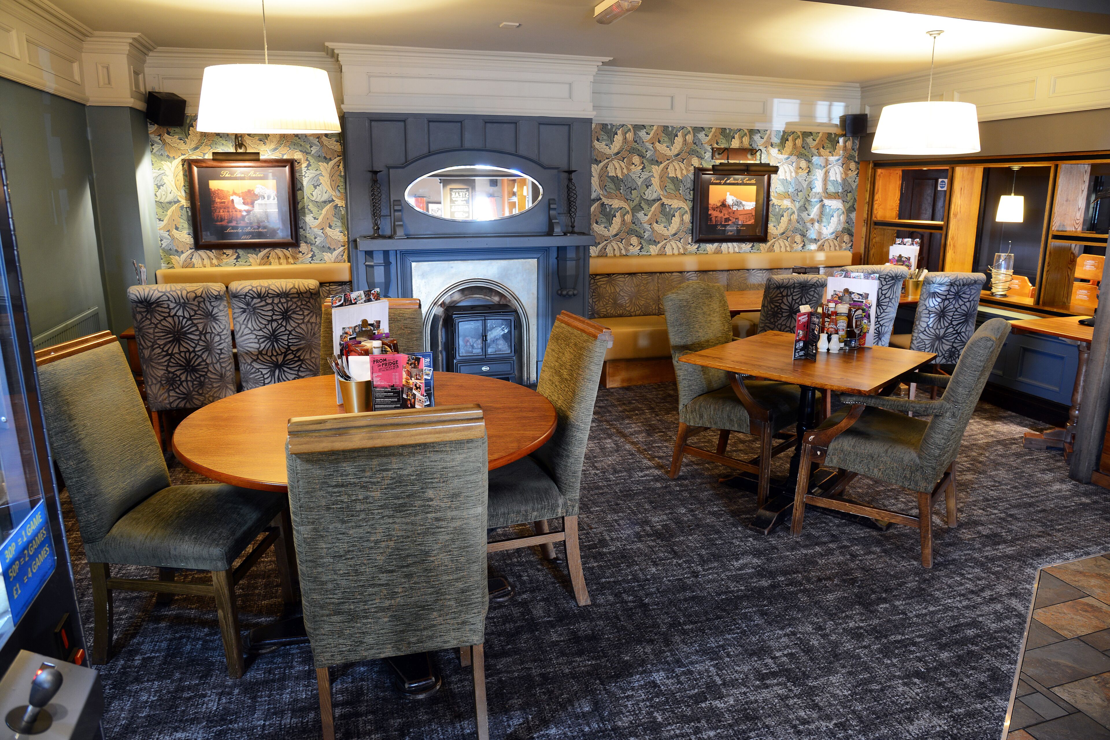 A hotel dining area with round tables and cushioned chairs, a fireplace, and patterned wallpaper.