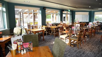 A restaurant with multiple wooden tables and chairs, a carpeted floor, and large windows allowing natural light.