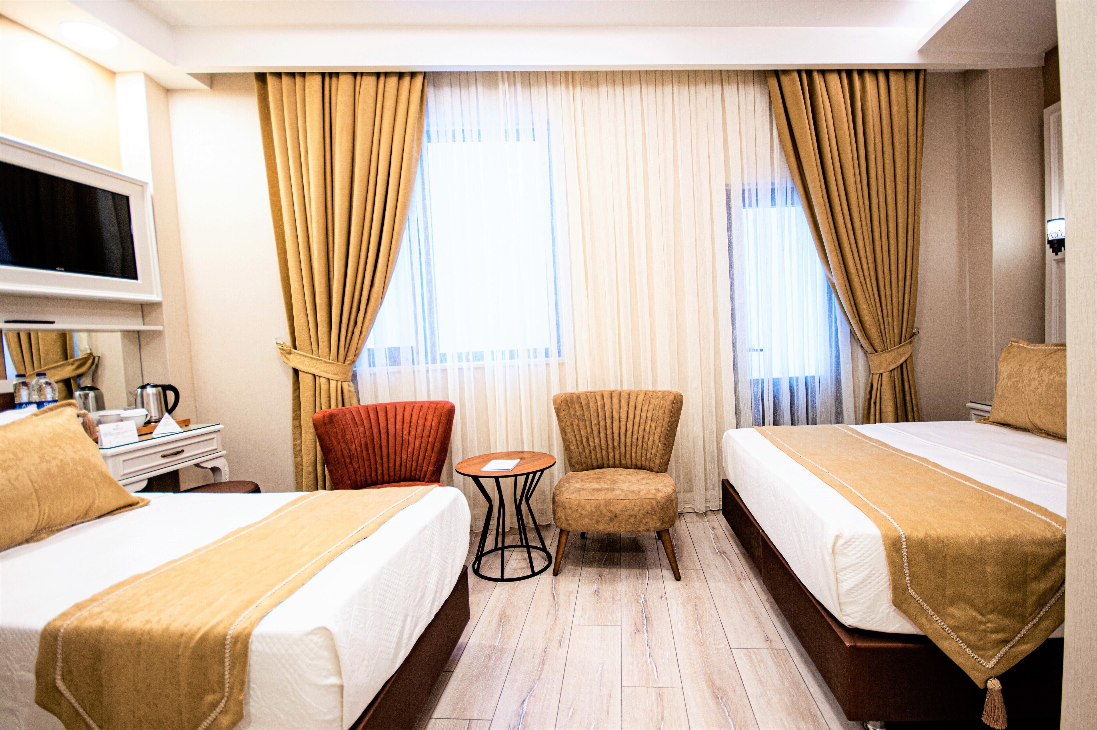 A hotel room with two beds, a TV, a desk with a kettle, and a chair.