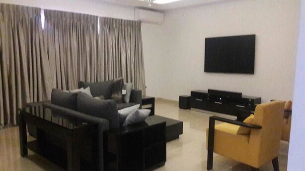 A modern living room with a sectional sofa, a yellow armchair, and a flat-screen TV mounted on the wall. - Koza Town (Lagos)