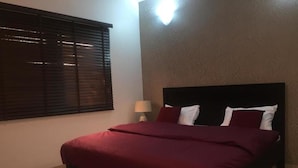 A bedroom with a bed, a window with blinds, a nightstand with a lamp, and a textured wall. - Koza Town (Lagos)
