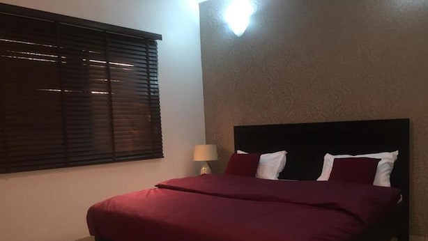 A bedroom with a bed, a window with blinds, a nightstand with a lamp, and a textured wall. - Koza Town (Lagos)