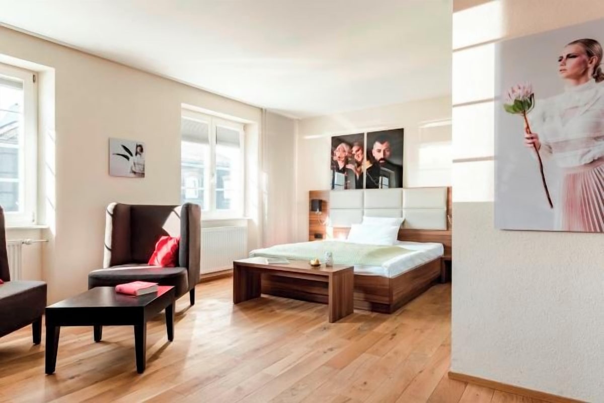 A modern hotel room with a large bed, a sitting area with a chair and a table, and artwork on the walls.