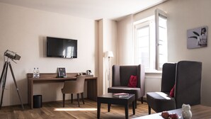 A modern living room with a wooden floor, a desk with a computer, a television, and a comfortable seating area with a sofa and armchairs. - Kunst-Hotel Drei König am Marktplatz Stadt Lörrach (Loerrach)