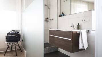 Un baño moderno con un tocador de madera, un lavabo blanco y un espejo sobre él. Hay un cabezal de ducha y una toalla colgada en la pared.
