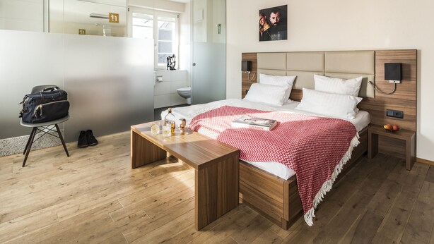 A modern hotel room with a wooden bed, a bedside table, a nightstand, a mirror, and a view of the bathroom. - Kunst-Hotel Drei König am Marktplatz Stadt Lörrach (Loerrach)