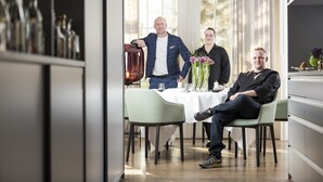 A modern dining area with a table set for four, a bar area with bottles, and a view through a large window. - Kunst-Hotel Drei König am Marktplatz Stadt Lörrach (Loerrach)