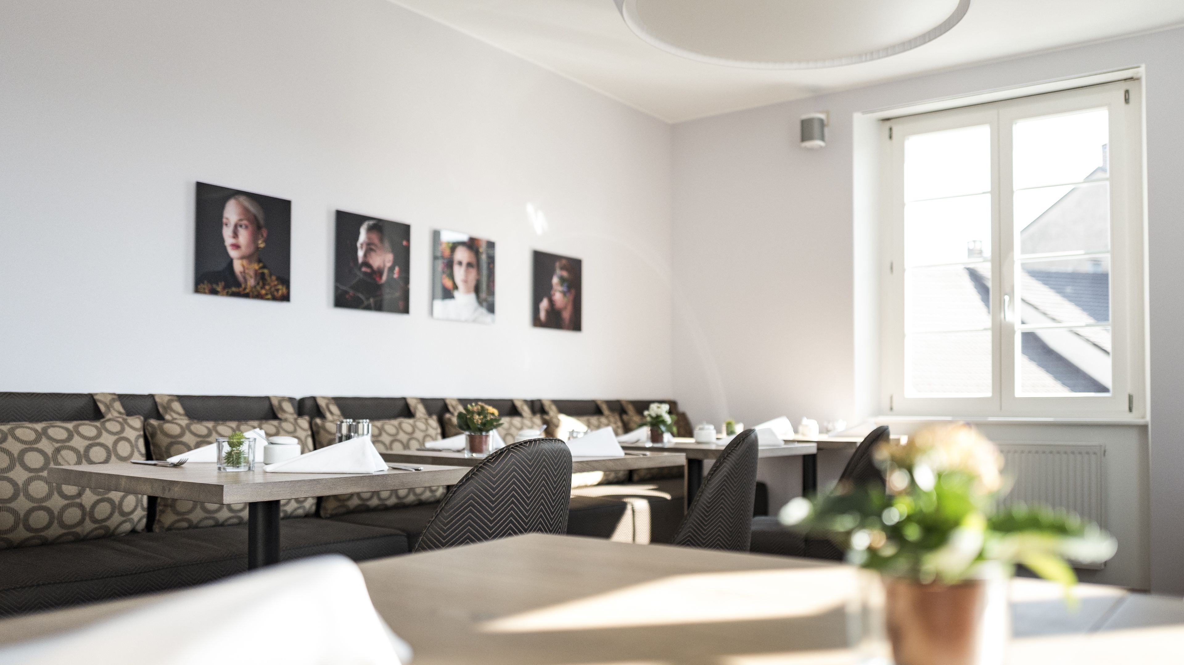 A modern dining area with a long bench, tables, and chairs. There are framed pictures on the wall and a window with a view of buildings.