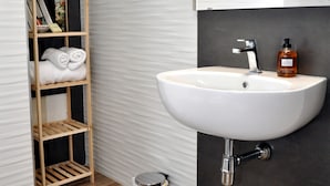 Un baño con lavamanos blanco, repisa de madera con toallas, un cubo de basura y un espejo.