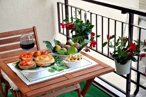 A wooden table with a white placemat, a glass of red wine, a plate of food including bread, tomatoes, and a pastry, and a bowl of nuts. There are potted plants with red flowers on a balcony railing. - B&B Kalurya (Lecce)