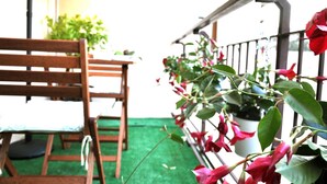 Una silla de madera y una pequeña mesa sobre una superficie verde, con una barandilla y macetas con flores rojas al fondo.