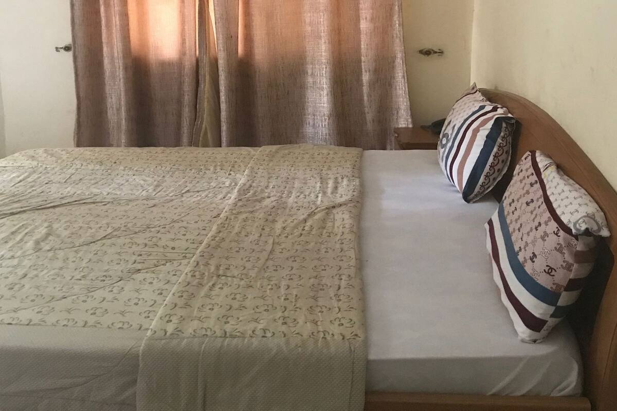 Cama com colcha estampada e almofadas, cabeceira de madeira e janela com cortinas.