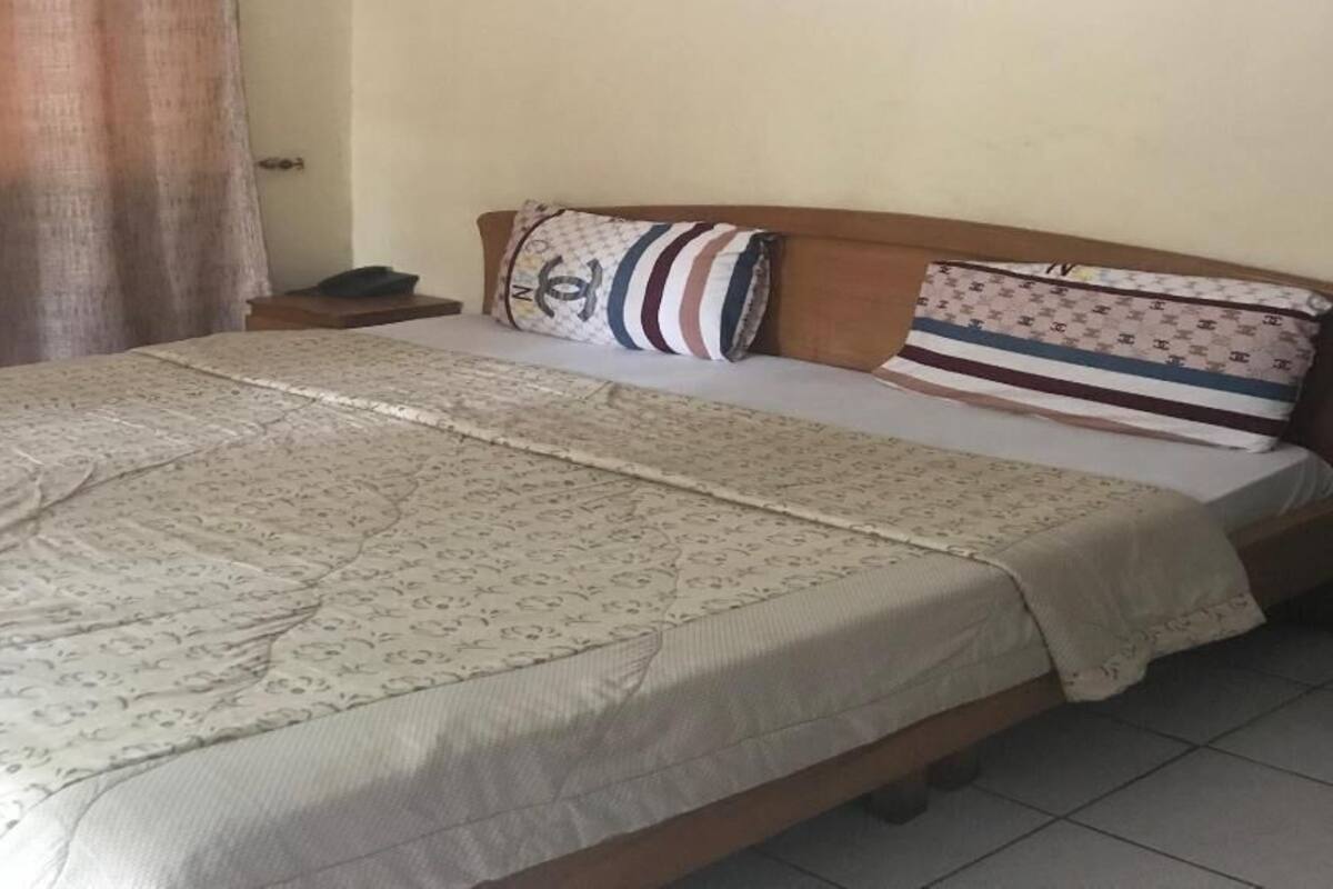 Uma cama com colcha estampada, duas almofadas com desenhos diferentes, uma mesinha de cabeceira com um telefone e uma cortina entreaberta.