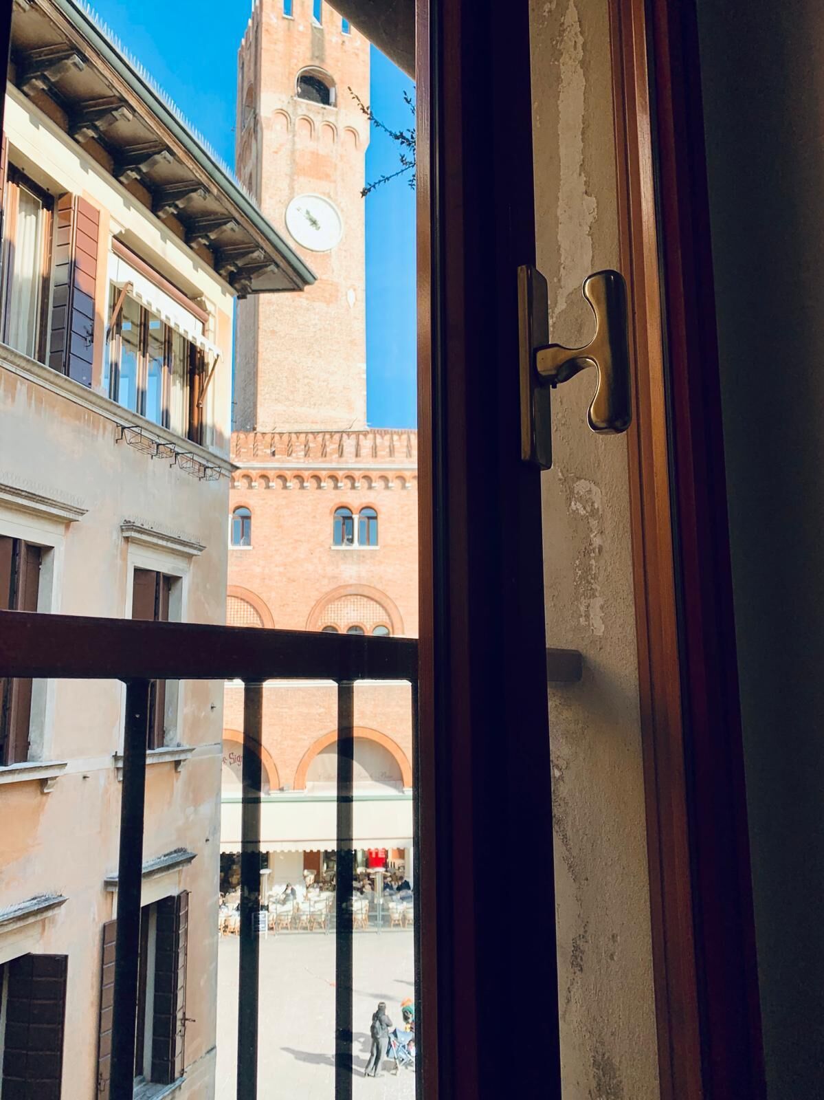 Blick durch ein Fenster auf ein Gebäude mit einem Uhrturm, Balkonen und einer Straße mit Außenplätzen.