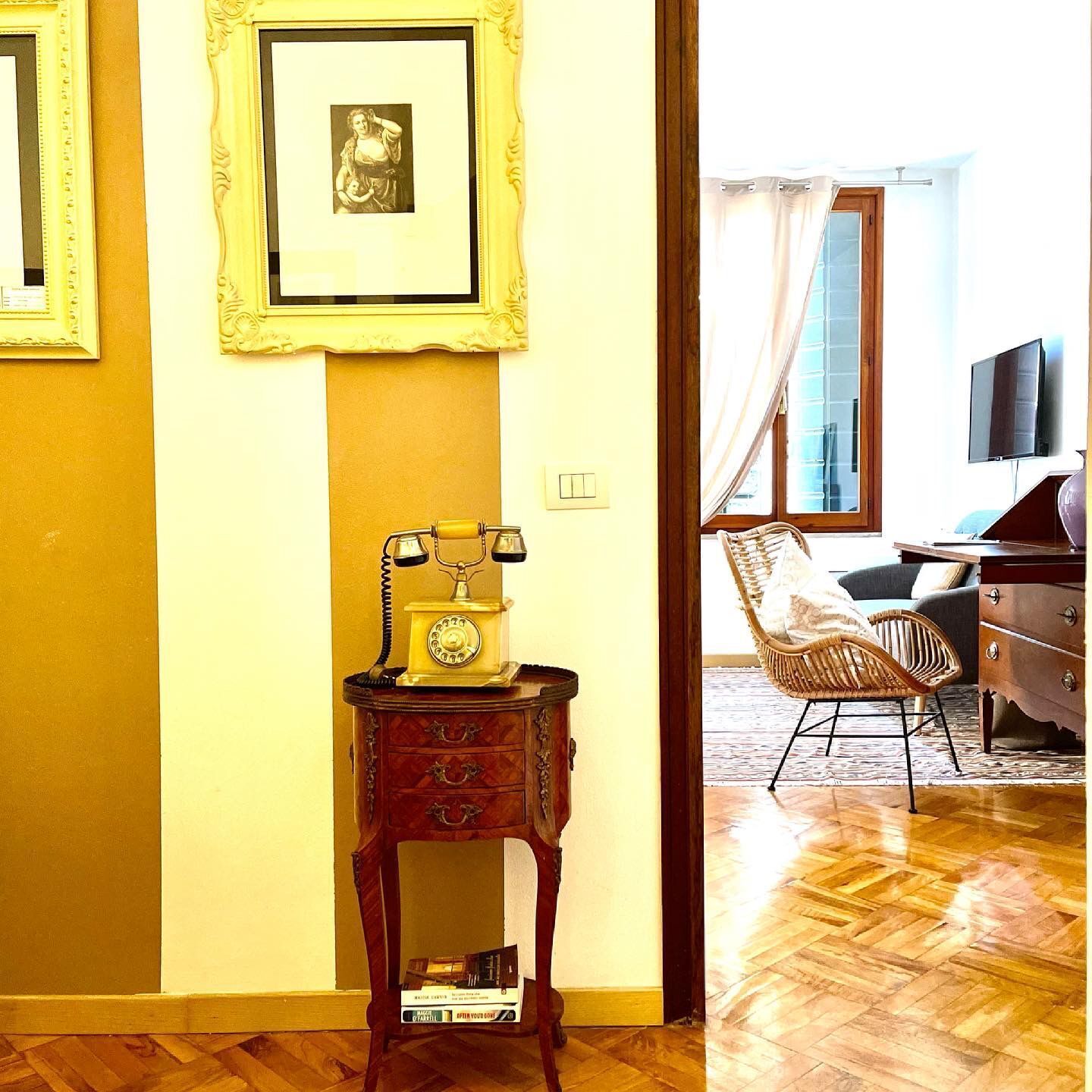 Een kamer met een houten vloer, een klein houten bijzettafeltje met een draaischijf telefoon, een ingelijst beeld en een rieten stoel.