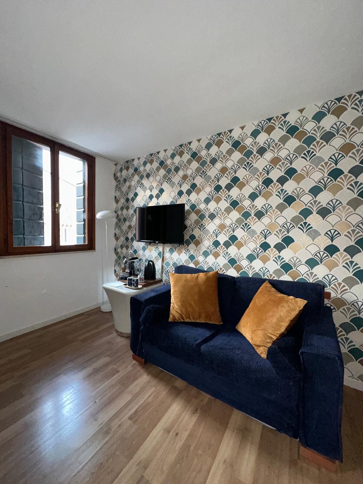 A room with a dark blue sofa, two mustard-colored pillows, a white side table with a lamp, and a patterned wallpaper.