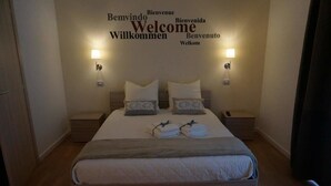 Een net opgemaakt bed met kussens en opgevouwen handdoeken, twee nachtlampjes en een muur met 'Welcome' in verschillende talen.