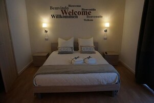 Una cama bien tendida con almohadas y toallas dobladas, dos lámparas de noche y una pared con la palabra 'Welcome' en varios idiomas.