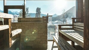 Un banco y una mesa de madera en una habitación con pared de piedra y ventanas amplias que dan a vistas de montañas y un paisaje nevado.