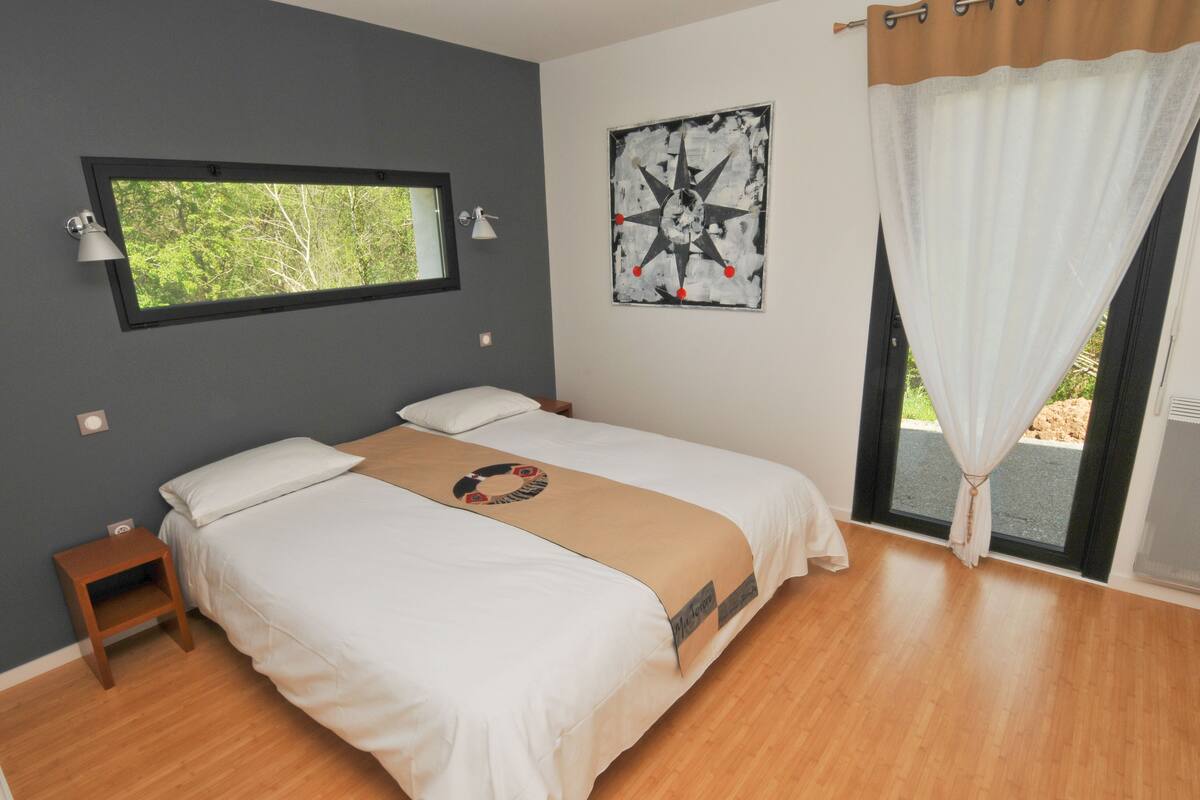 Une chambre à coucher avec un lit, une fenêtre, une lampe fixée au mur, un ventilateur de plafond et une vue sur la verdure à l’extérieur.