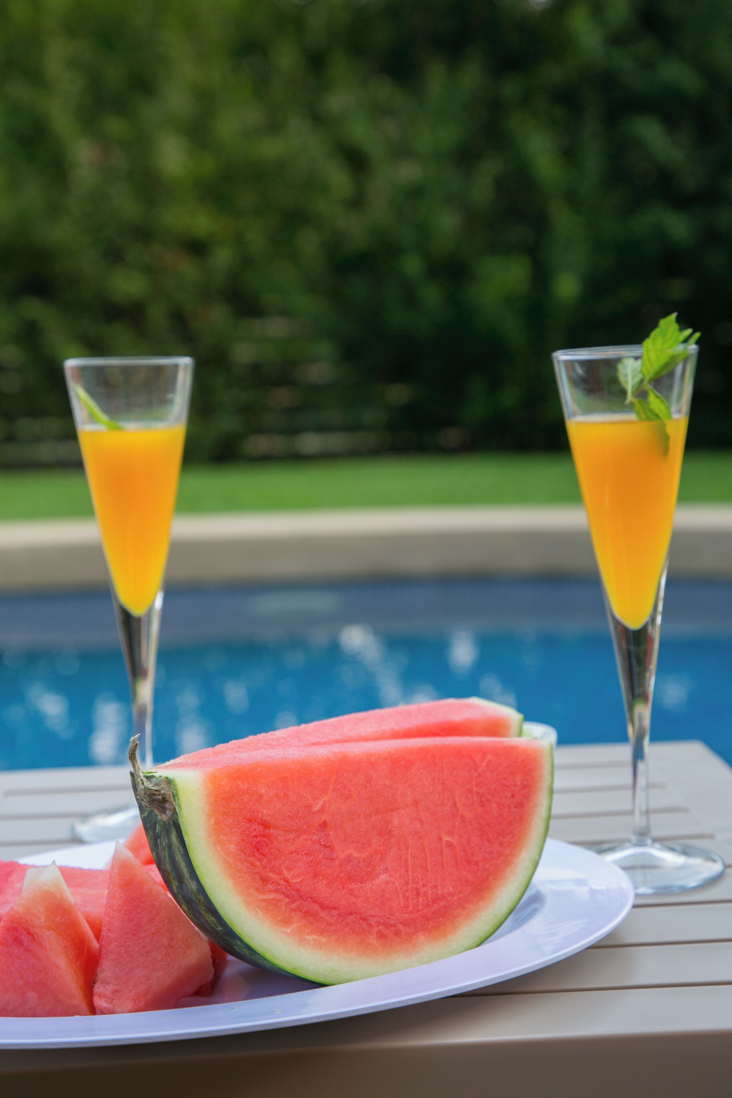 Mesa junto a la piscina con una fuente de sandía y dos vasos de jugo de naranja.