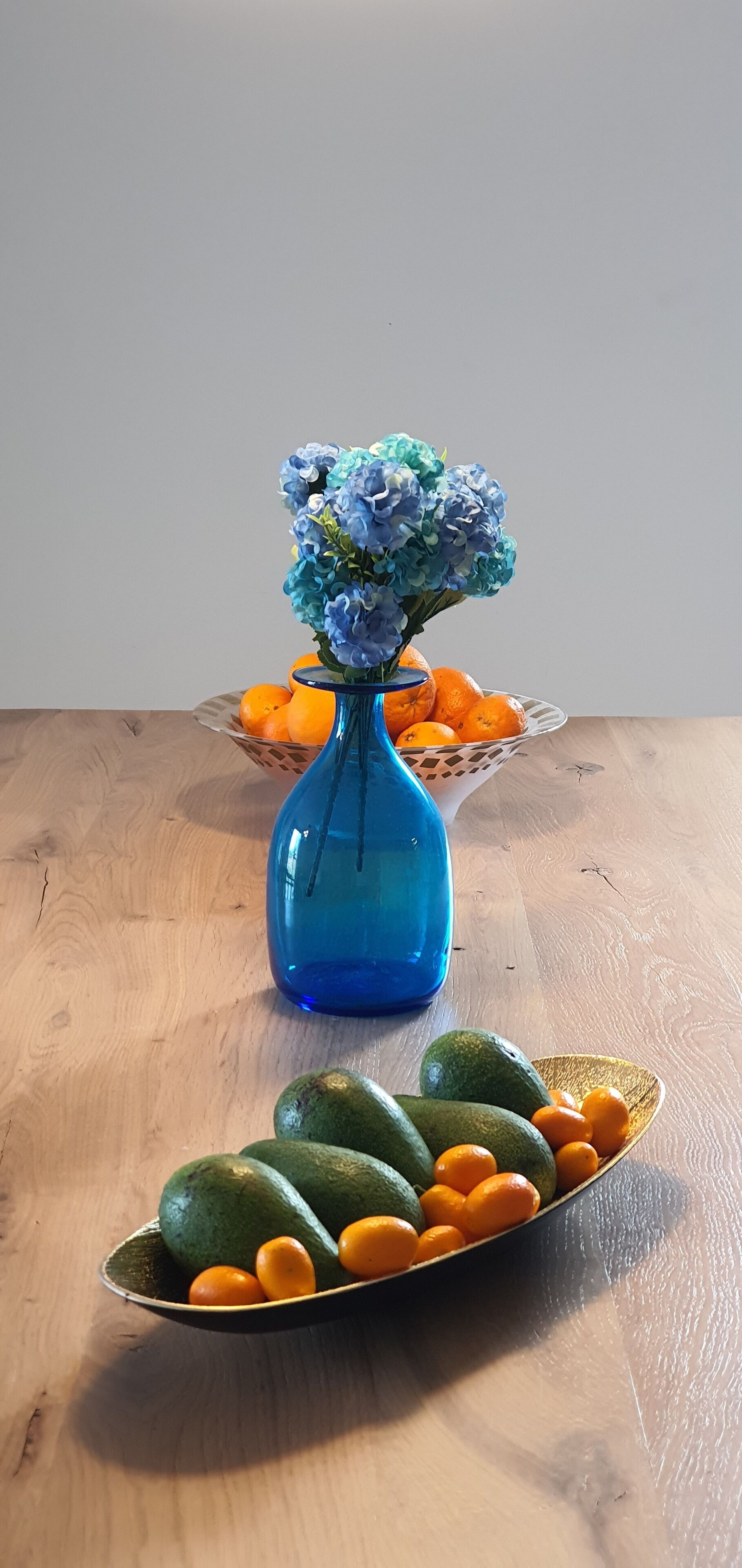 Ett träbord med en blå vas som innehåller blå blommor, en korg med apelsiner och en tallrik med avokado och apelsiner.