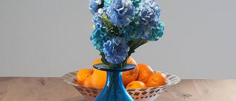 Ett träbord med en blå vas som innehåller blå blommor, en korg med apelsiner och en tallrik med avokado och apelsiner.