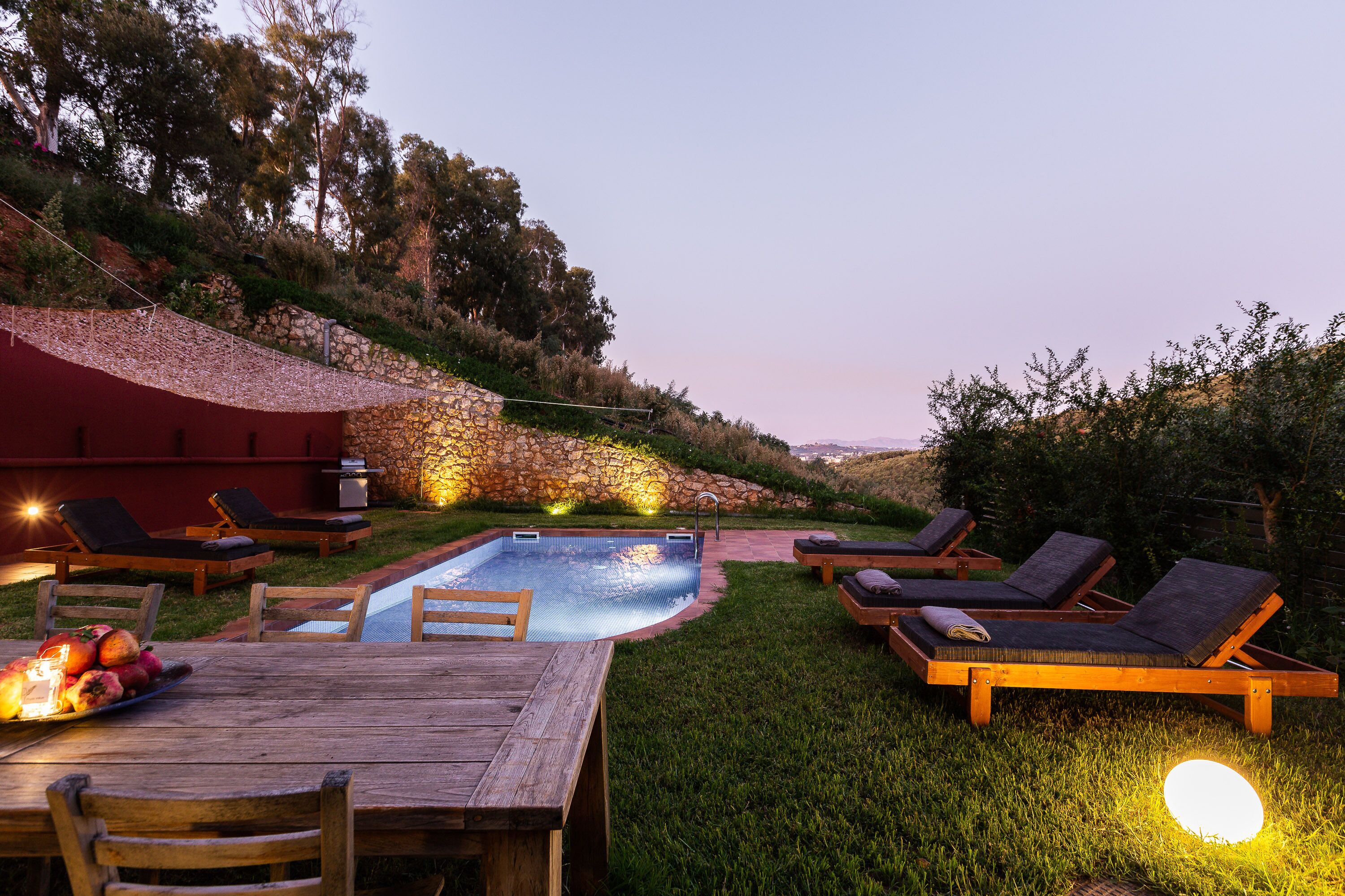 A poolside area with lounge chairs, a wooden table, and a stone wall.