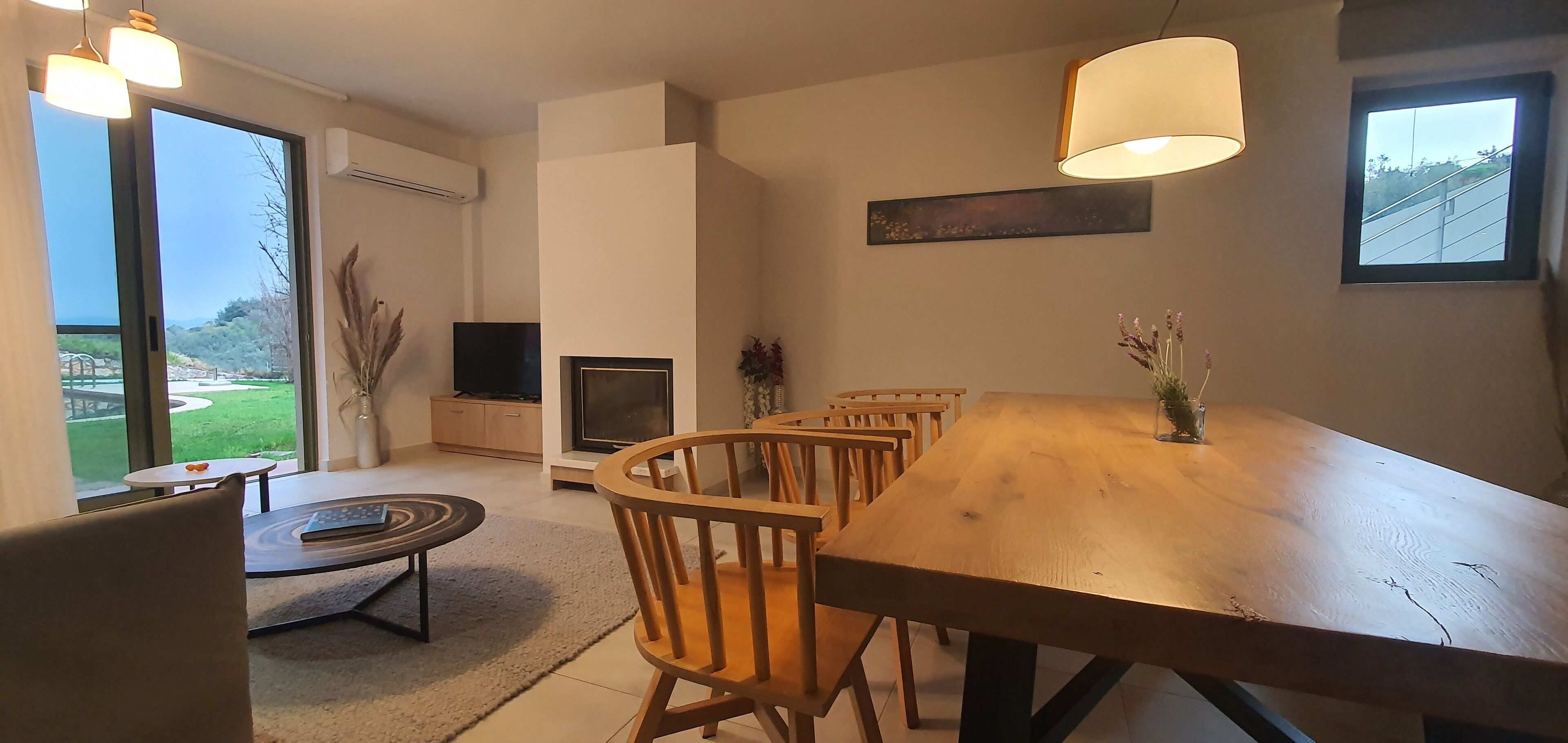 Un comedor con una mesa y sillas de madera, un televisor y vistas a un jardín a través de amplios ventanales.