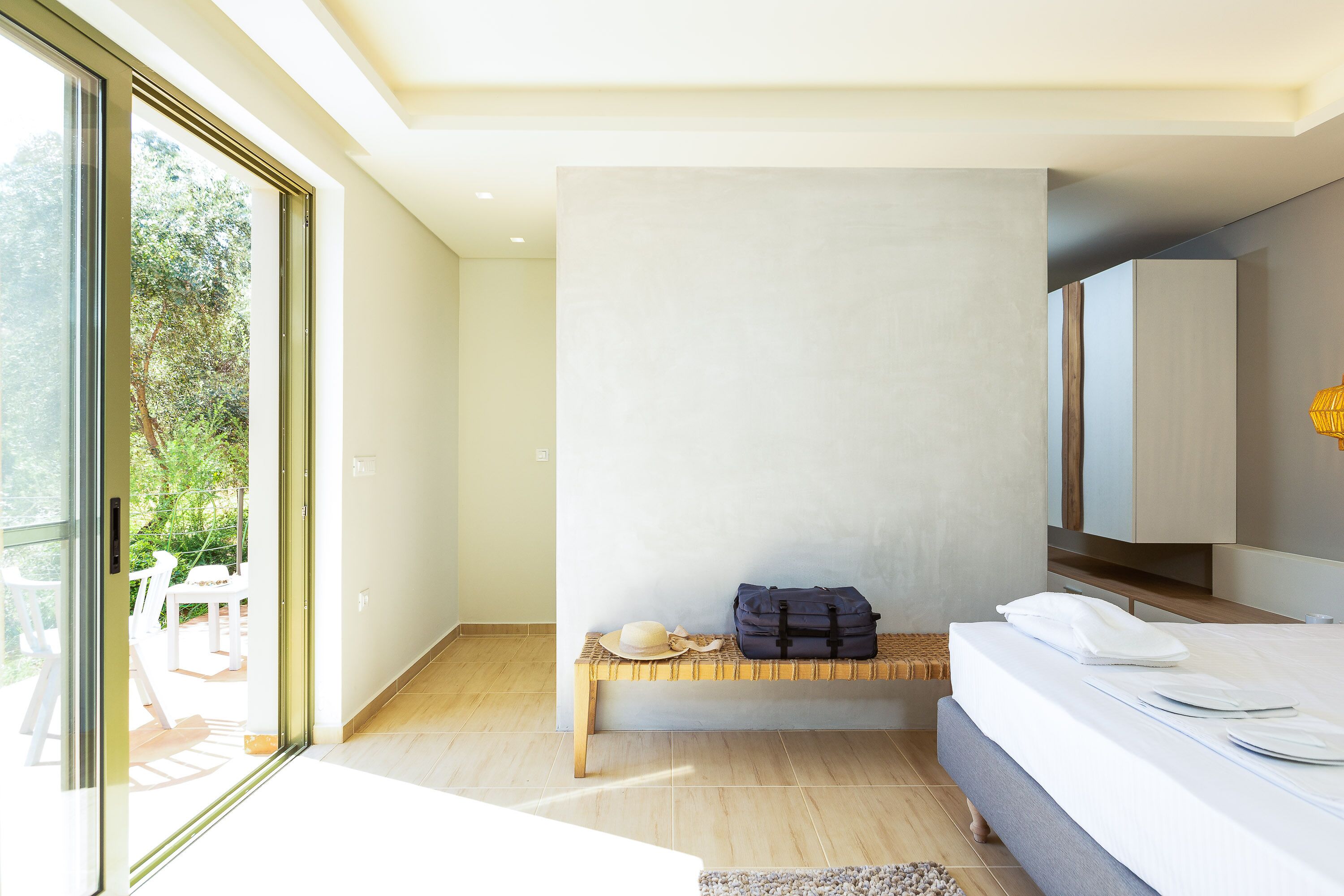 A modern bedroom with a large bed, a bench with luggage, and a view of greenery outside.