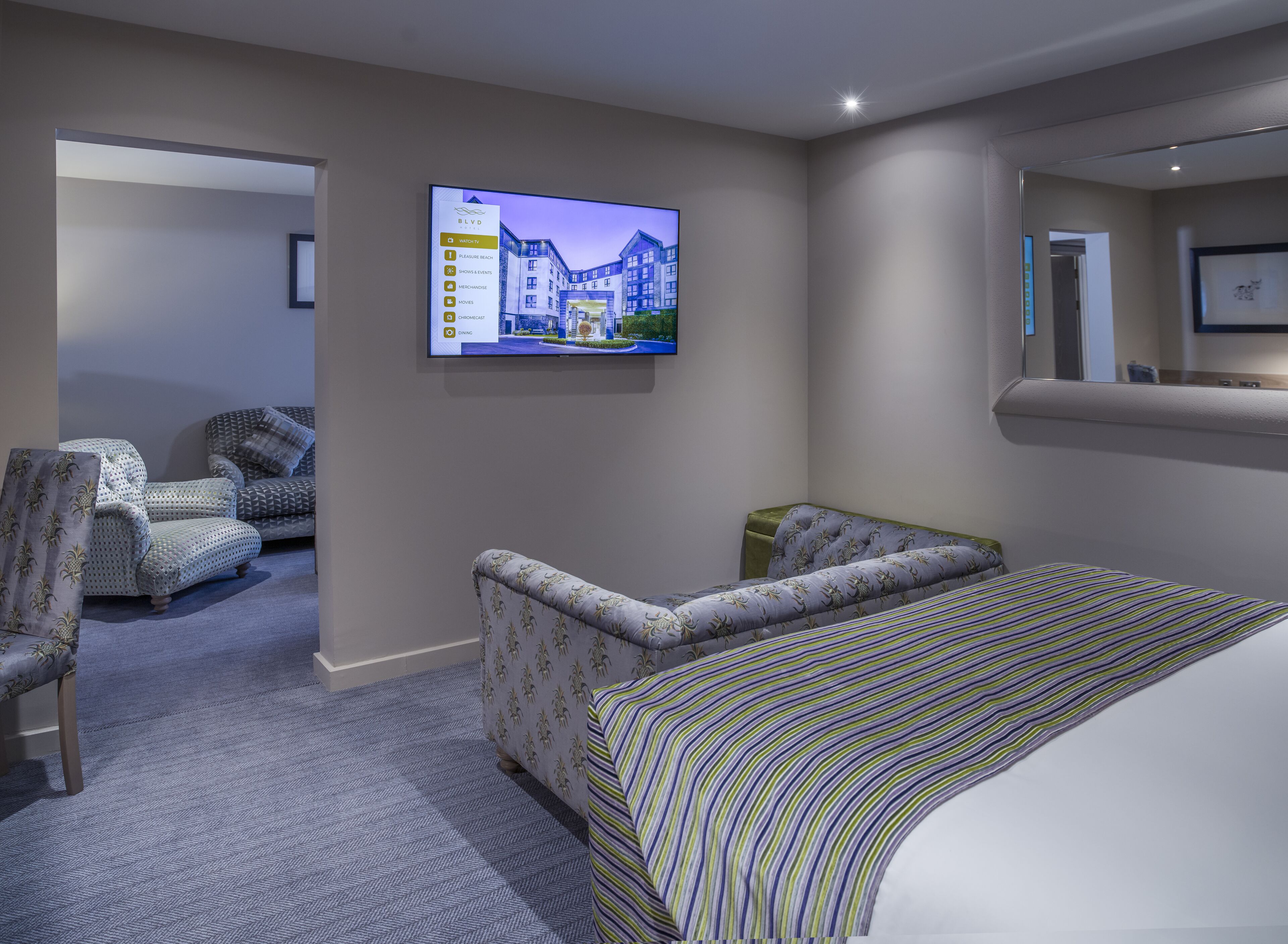 A hotel room with a large bed, two patterned armchairs, a television displaying a building image, and a carpeted floor.