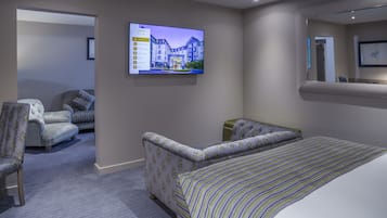 A hotel room with a large bed, two patterned armchairs, a television displaying a building image, and a carpeted floor.