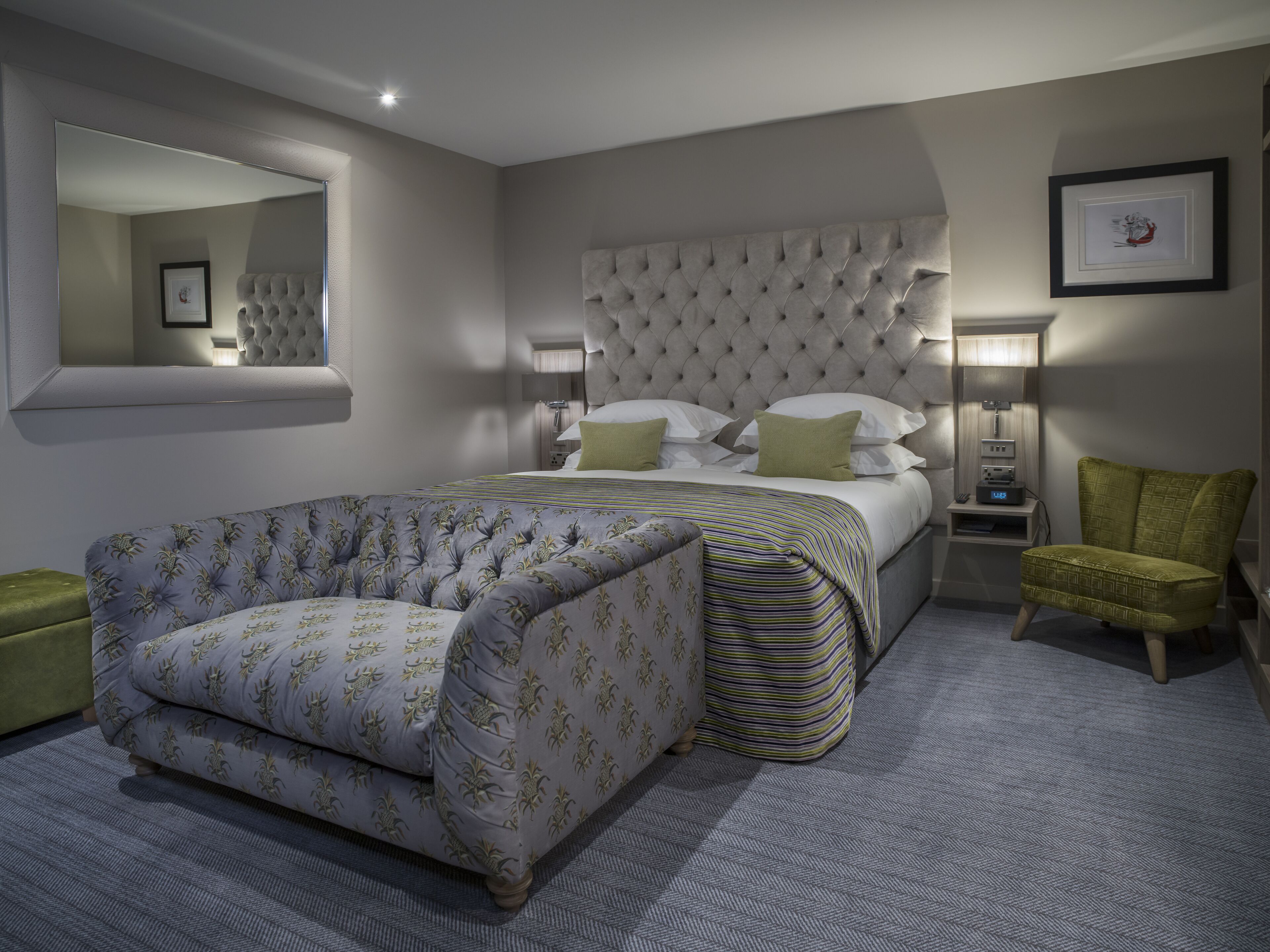 A bedroom with a tufted headboard, a large bed with pillows, a green armchair, a footstool, a bedside table with a lamp, and a wall-mounted mirror.