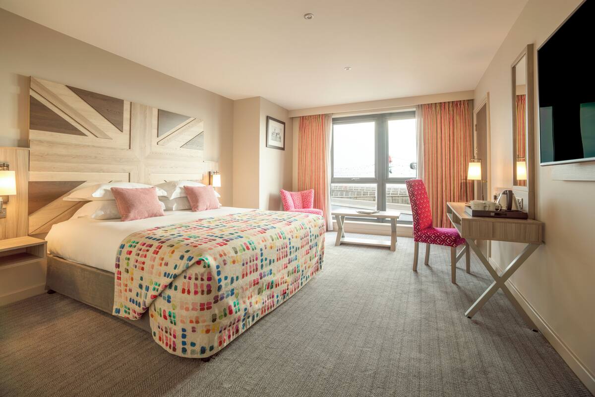 A hotel room with a bed, a desk, a chair, a large window, and a mirror.